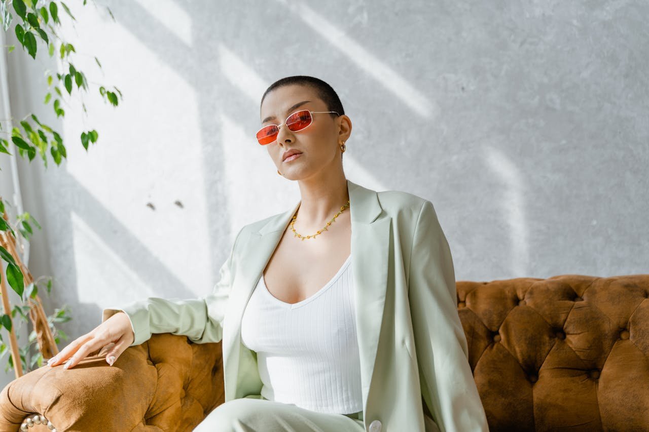 Confident woman in red sunglasses and chic suit sitting on a vintage couch in a sunlit room.